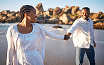 Loving young couple having romantic moment while holding hands and dancing or following each other while the wife takes lead at the beach. Happy african american lovers being playful and enjoying time by the sea