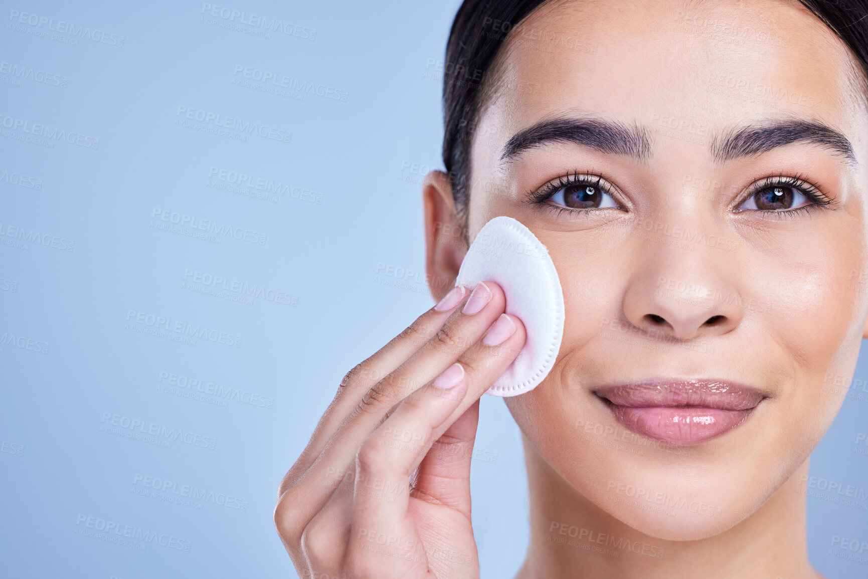 Buy stock photo Studio Portrait of a beautiful mixed race woman using a cotton pad to remove makeup during a selfcare grooming routine. Hispanic woman applying cleanser to her face against blue copyspace background