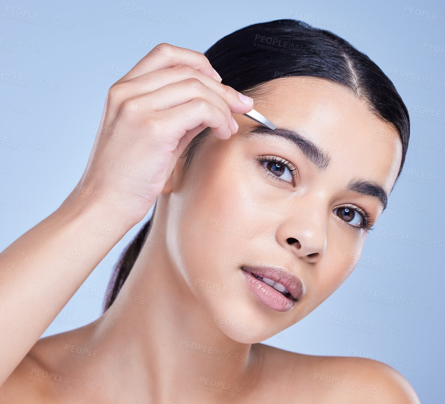 Buy stock photo Studio portrait of a beautiful mixed race young woman with glowing skin posing against blue copyspace background while tweezing her eyebrows. Hispanic model using a tweezer for hair removal