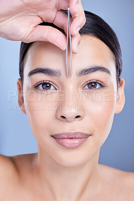 Studio portrait of a smiling mixed race young woman with glowing skin posing against blue copyspace background while tweezing her eyebrows. Hispanic model using a tweezer for hair removal