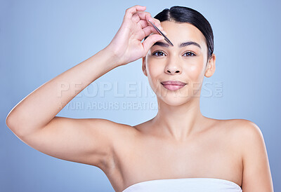 Studio portrait of a smiling mixed race young woman with glowing skin posing against blue copyspace background while tweezing her eyebrows. Hispanic model using a tweezer for hair removal