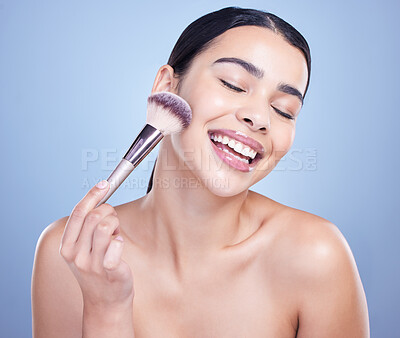 A smiling beautiful mixed race woman posing with a makeup brush during a pamper routine. Hispanic model holding a contouring brush against a blue copyspace background