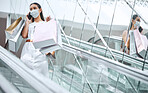One young mixed race woman wearing a medical face mask for prevention against coronavirus and talking on a cellphone while on an escalator after a shopping spree. Hispanic woman carrying retail bags in a mall during Covid-19 pandemic