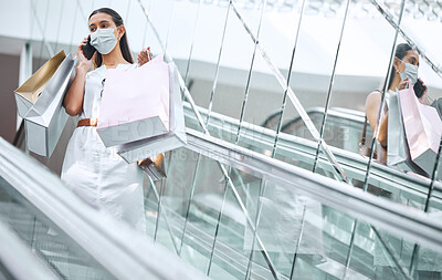 Buy stock photo One young mixed race woman wearing a medical face mask for prevention against coronavirus and talking on a cellphone while on an escalator after a shopping spree. Hispanic woman carrying retail bags in a mall during Covid-19 pandemic