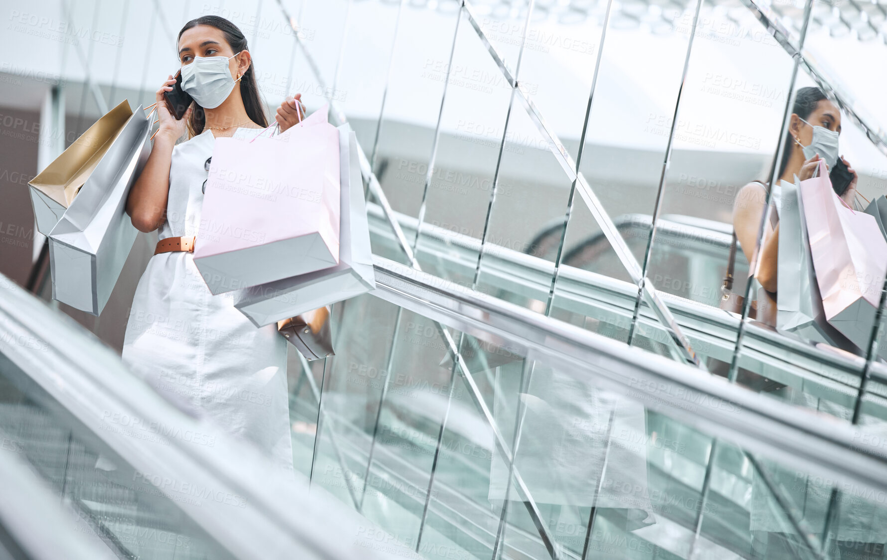 Buy stock photo One young mixed race woman wearing a medical face mask for prevention against coronavirus and talking on a cellphone while on an escalator after a shopping spree. Hispanic woman carrying retail bags in a mall during Covid-19 pandemic