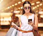 Stylish young woman wearing sunglasses using a cell phone while carrying bags during a shopping spree. Young brunette woman sending a text with her smartphone after shopping in a mall. Woman checking sale offers online or using discount app