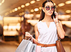 A young stylish woman wearing sunglasses and talking on cellphone while carrying bags during a shopping spree. Young brunette woman on a call with her smartphone while shopping in a mall