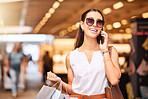 A happy young mixed race woman wearing sunglasses using a cell phone while carrying bags during a shopping spree. Young brunette woman on a call with her smartphone while enjoying shopping in a mall