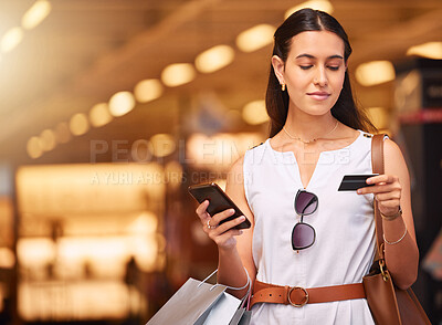A young mixed race woman using a cell phone and bank card while carrying bags during a shopping spree. Young brunette woman purchasing items online with her smartphone and credit card in a shopping mall. Woman using banking app to increase spending limit