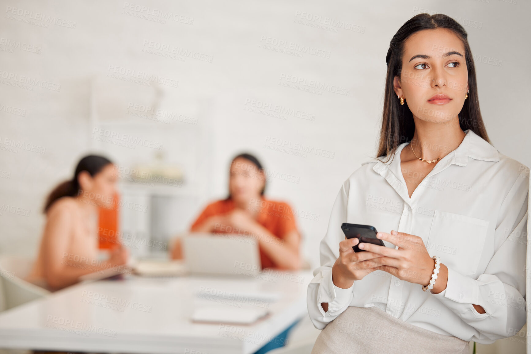Buy stock photo One young hispanic business woman thinking while texting on a cellphone in an office with her colleagues in the background. Entrepreneur making a choice and decision while browsing and planning online with smartphone