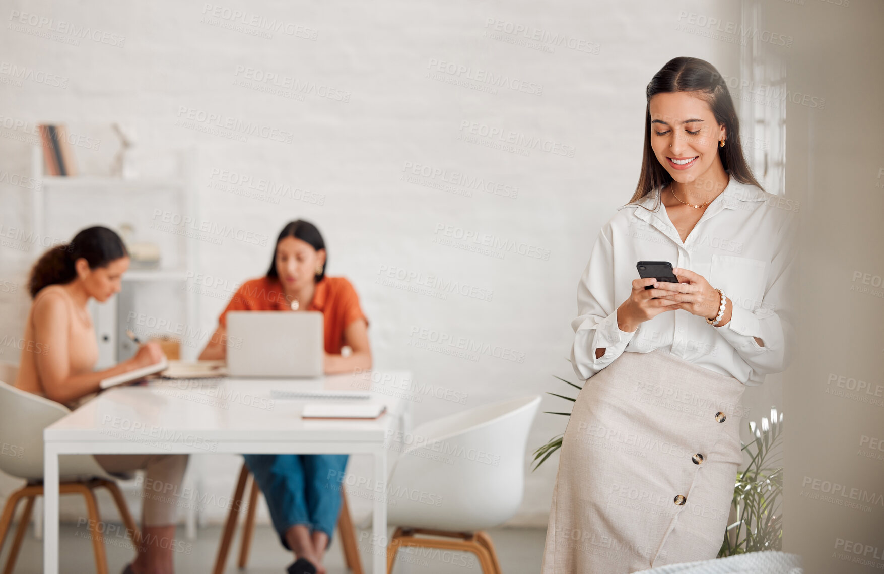 Buy stock photo One happy young hispanic business woman texting on a cellphone in an office with her colleagues in the background. Confident entrepreneur browsing and planning online with smartphone