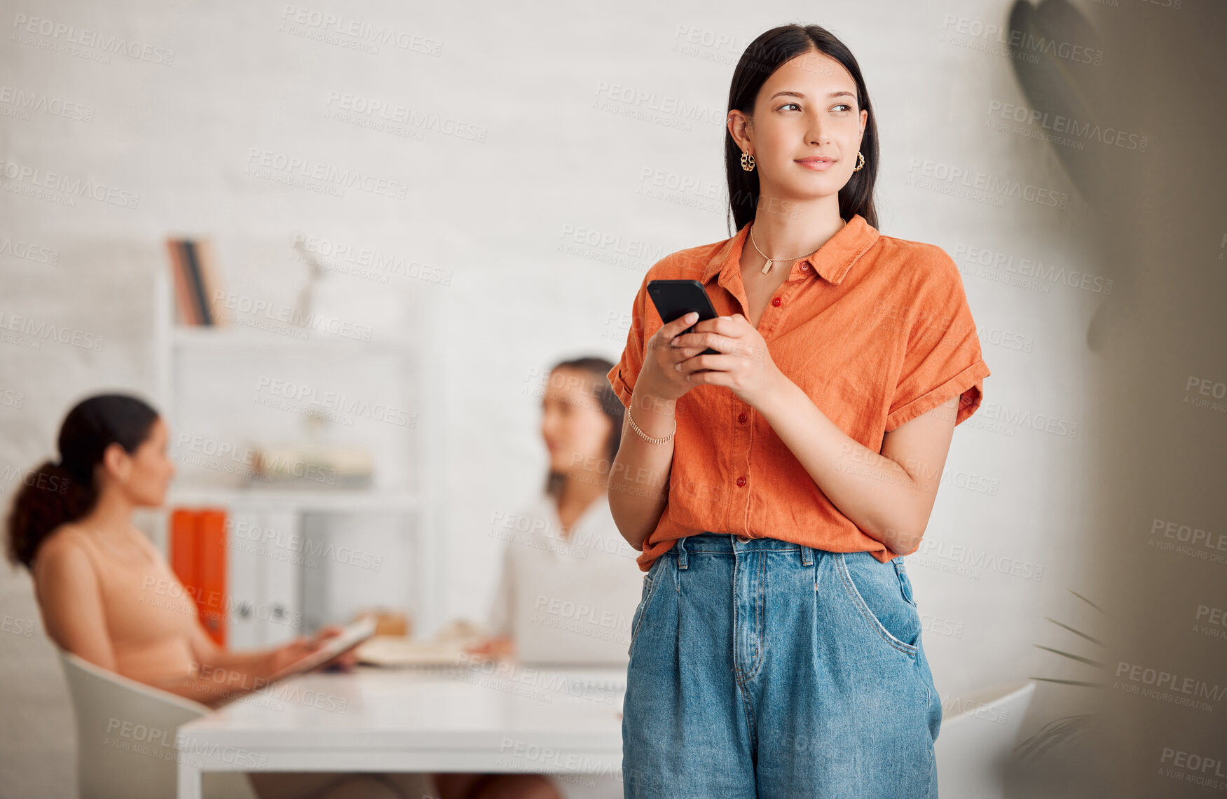 Buy stock photo One young hispanic business woman thinking while texting on a cellphone in an office with her colleagues in the background. Entrepreneur making a choice and decision while browsing and planning online with smartphone