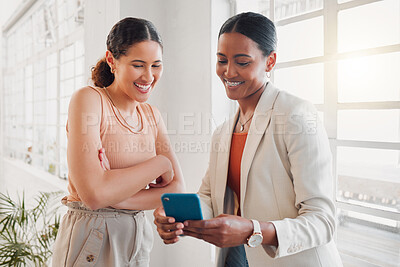 Two young happy mixed race businesswomen using a phone together at work. Hispanic businesspeople talking using social media on a cellphone together in an office