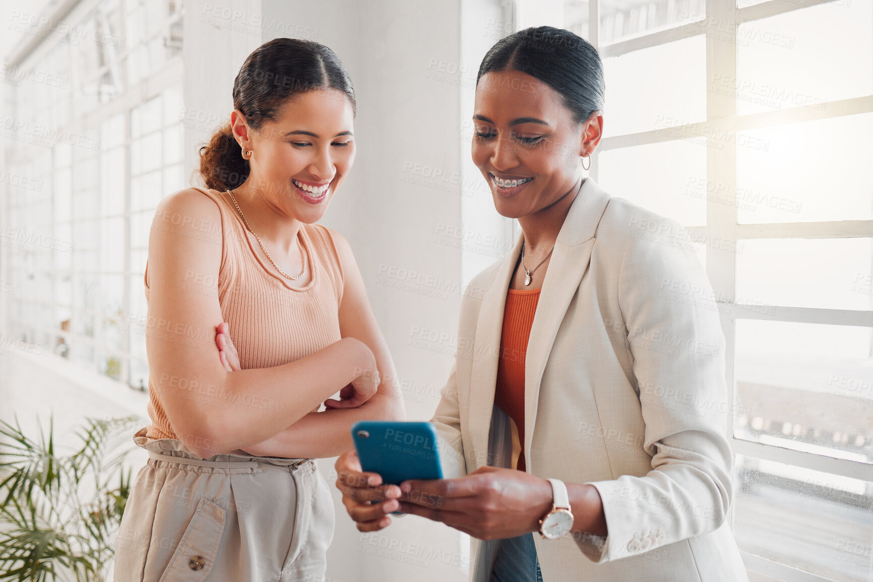 Buy stock photo Two young happy mixed race businesswomen using a phone together at work. Hispanic businesspeople talking using social media on a cellphone together in an office