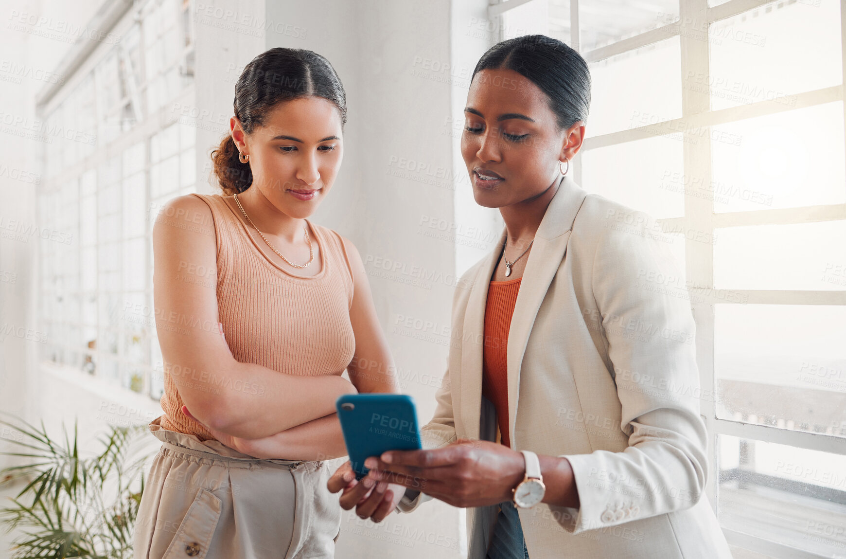 Buy stock photo Two young happy mixed race businesswomen using a phone together at work. Hispanic businessperson talking while showing a female colleague her cellphone in an office