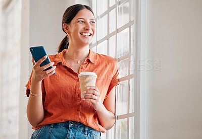Young happy mixed race businesswoman using her phone while drinking a coffee and looking out of a window alone in an office at work. One cheerful hispanic businessperson using social media on her cellphone while holding a coffee cup and thinking on a brea