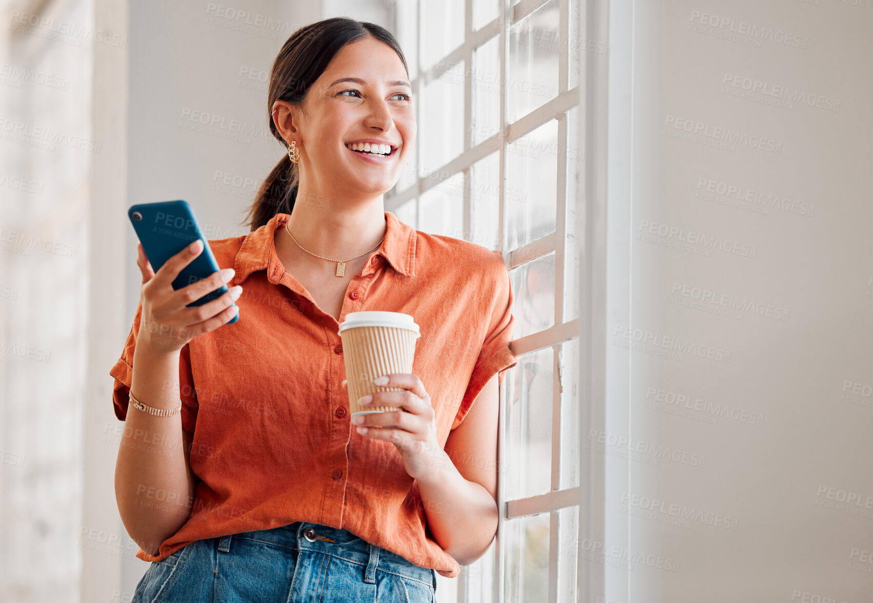 Buy stock photo Young happy mixed race businesswoman using her phone while drinking a coffee and looking out of a window alone in an office at work. One cheerful hispanic businessperson using social media on her cellphone while holding a coffee cup and thinking on a break while standing at work