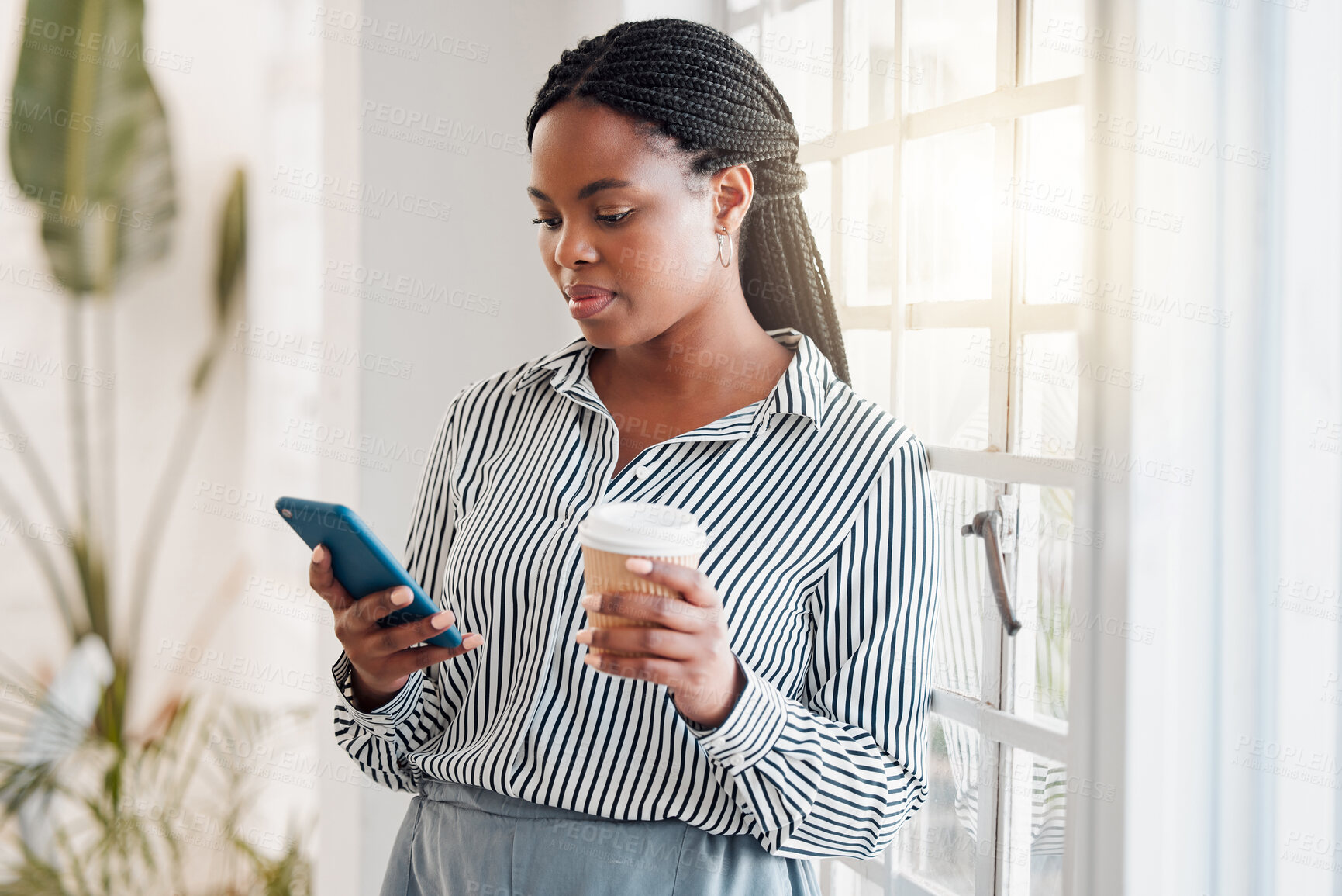 Buy stock photo Young african american businesswoman using her phone while drinking a coffee alone in an office at work. One black woman using social media on her cellphone and holding a coffee cup on a break standing at work