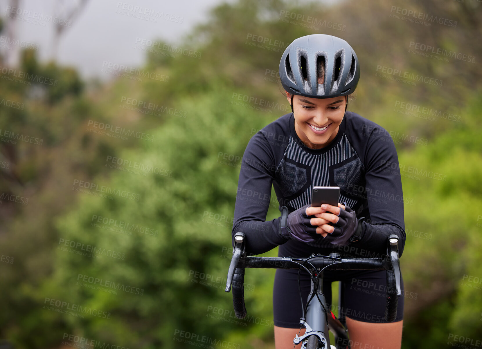 Buy stock photo One athletic young woman using a cellphone while cycling outside on a bicycle. Sporty fit female wearing a helmet and taking a break to text, browse online social media and use a navigation app
