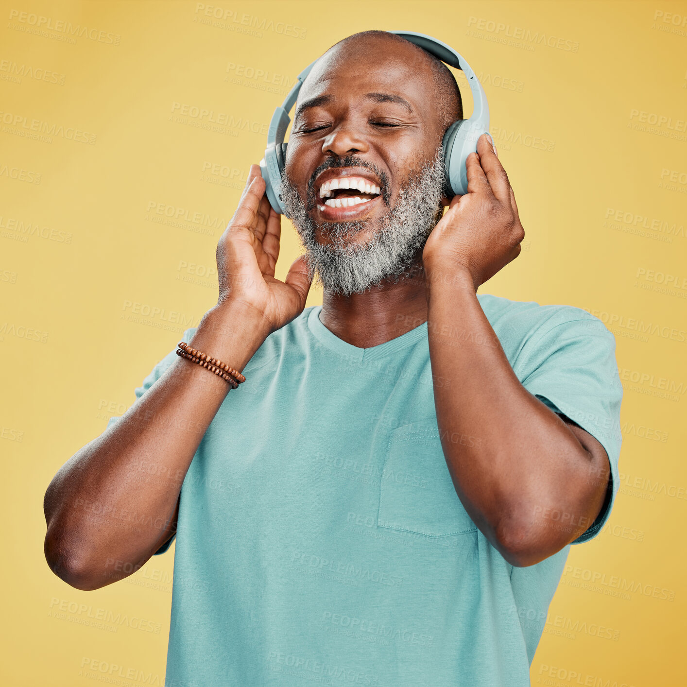 Buy stock photo Happy mature African American man standing alone against a yellow background in a studio and wearing headphones to listen to music. Smiling portrait of senior black man with grey beard enjoying music