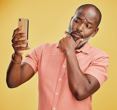 One contemplative trendy mature African American man taking selfies on a cellphone against a yellow studio background. Fashionable black man standing and posing while taking pictures for social media