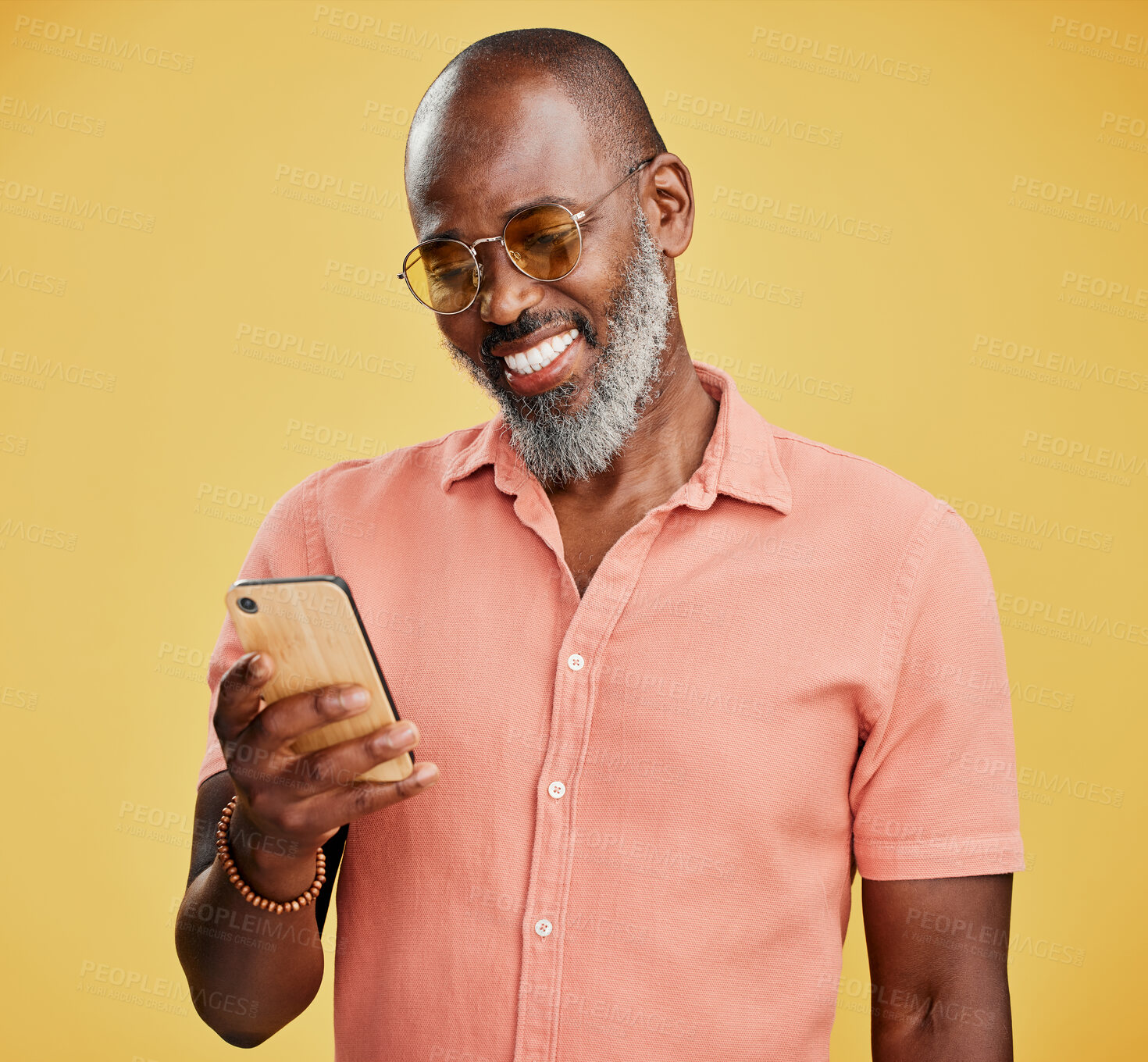 Buy stock photo One mature african american using his phone while standing in studio isolated against a yellow background. Handsome man with a beard and wearing glasses reading and sending text message on his mobile