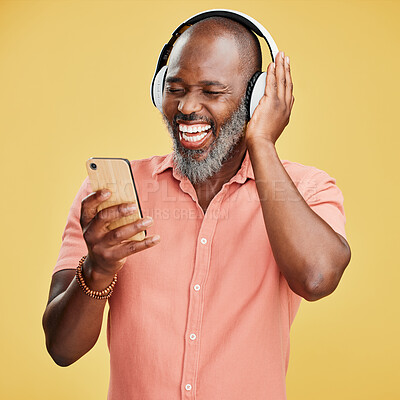 One mature african american man listening to music using wireless headphones while isolated against a yellow background. Happy man with a grey beard smiling while streaming on his phone in studio