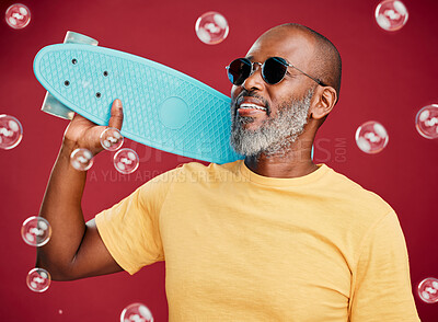 One mature african american man standing with a mini skateboard surround by many bubbles in studio isolated against a red background. Handsome and carefree man wearing sunglasses and smiling happily