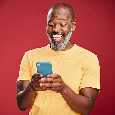 One mature african american using his phone while standing in studio isolated against a red background. Handsome man with a beard reading and sending text message on his mobile while laughing happily