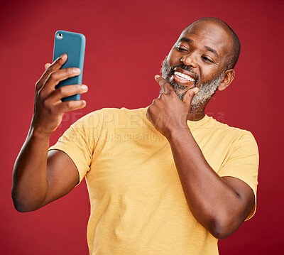 One contemplative trendy mature african american man taking a selfie on a smart phone against a red studio background. Fashionable black man standing and posing while taking pictures for social media