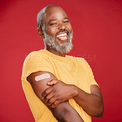 Buy stock photo Mature african american man with a beard smiling and looking happy while wearing a bandaid and holding his arm and standing against a red studio background. Vaccination, health and healthcare