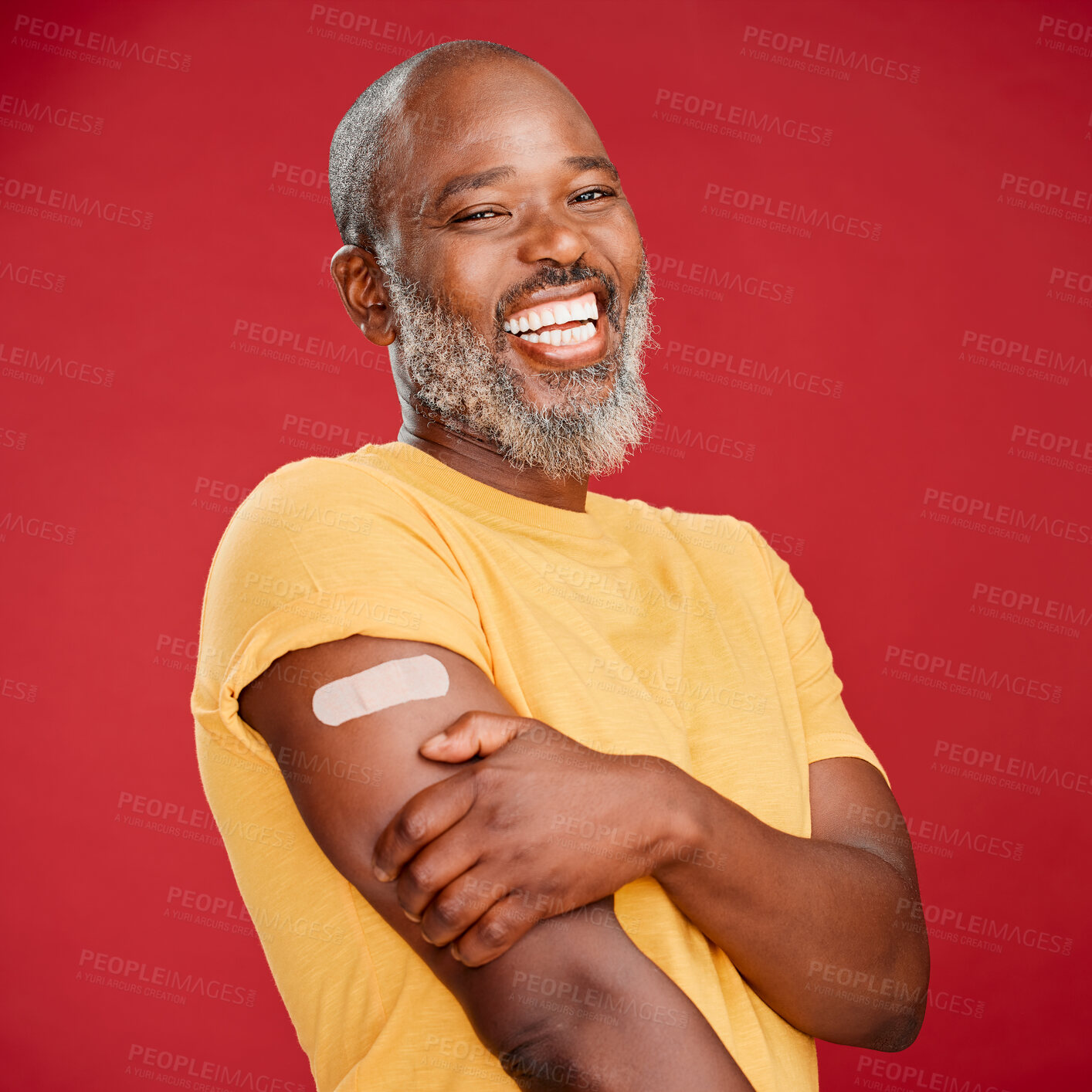 Buy stock photo Mature african american man with a beard smiling and looking happy while wearing a bandaid and holding his arm and standing against a red studio background. Vaccination, health and healthcare
