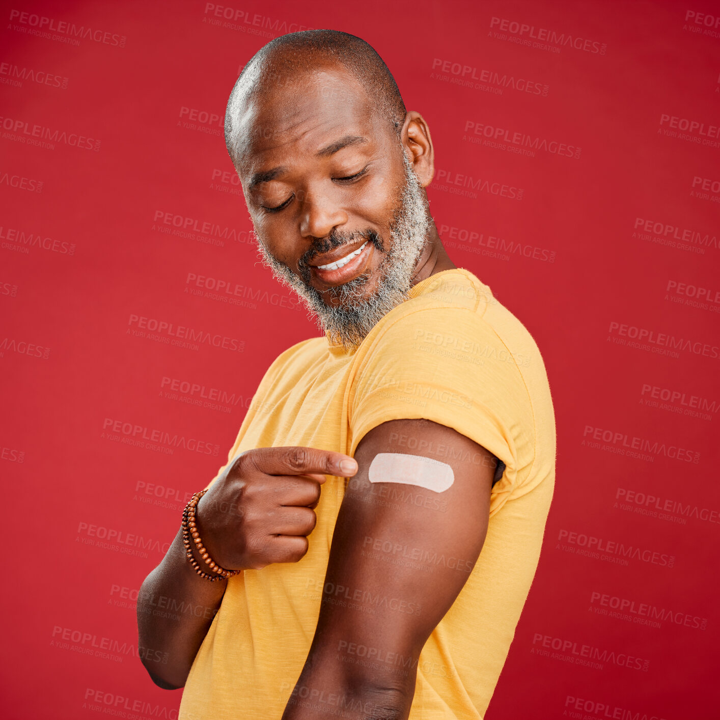 Buy stock photo A mature african american male pointing at a plaster on his arm against a red studio background. Black african man smiling after a minor injury and  making a hand gesture towards the plaster on his arm. 
