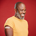 Mature african american man with a beard smiling and looking happy while wearing a bandaid and holding his arm and standing against a red studio background. Vaccination, health and healthcare