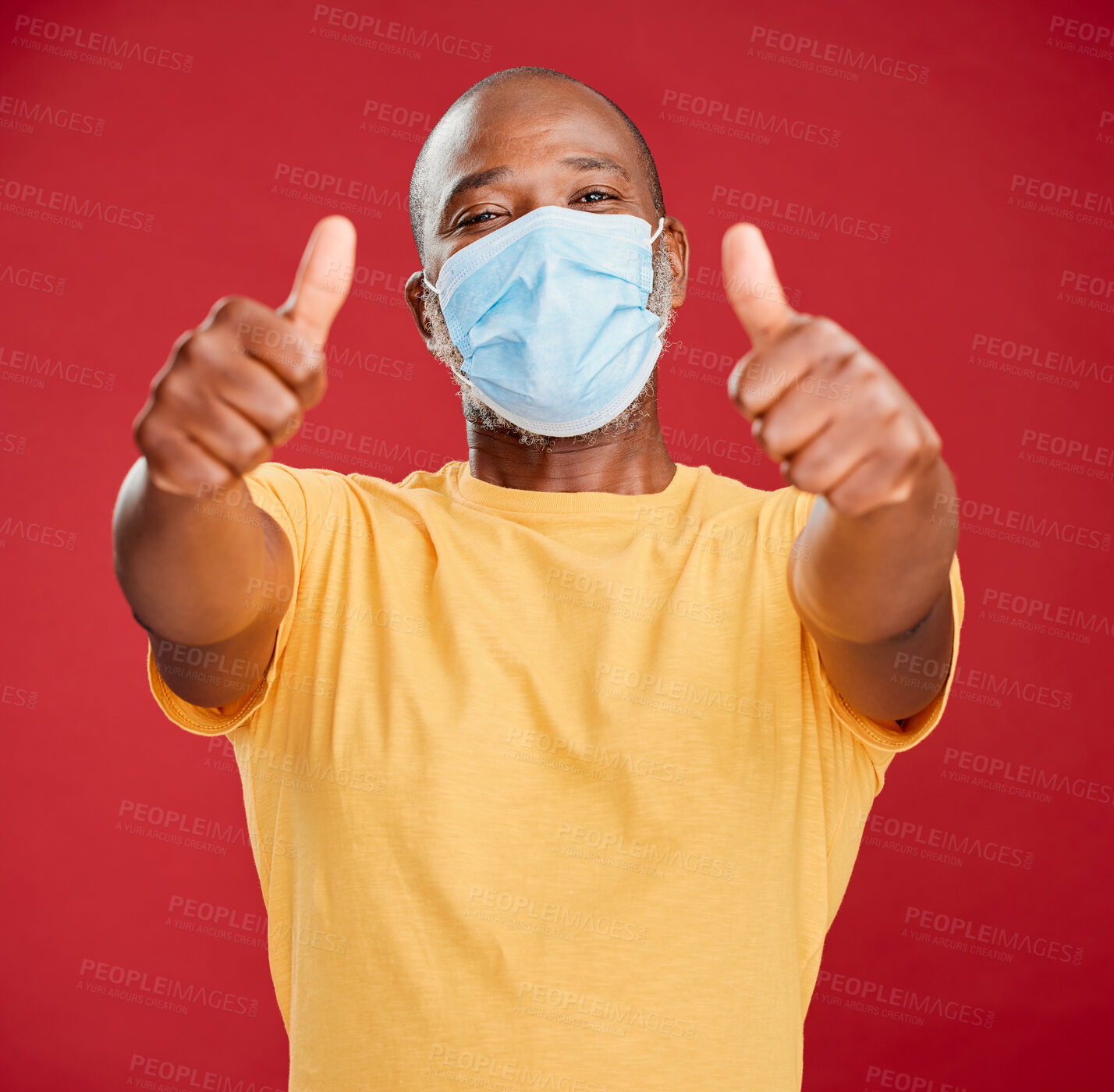 Buy stock photo A african american making wearing mask to stop the spread of coronavirus and standing against a red studio background while showing a thumbs up gesture with his hand. Black male suggesting that everything will be alright. Look after our health wearing a mask is great