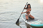 Young active african female smiling and kneeling on her paddle board on a lake out in nature. Young female enjoying a summer day at dawn on a paddle board. Life is better when you’re paddling in the water