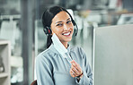 Young female call center agent smiling and wearing a headset and mask working on a computer in an office at work. Customer service, support and sales. Giving advice and helping. Answering calls
