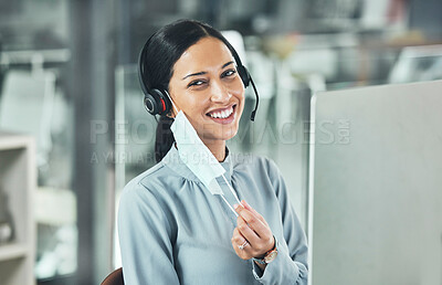 Young female call center agent smiling and wearing a headset and mask working on a computer in an office at work. Customer service, support and sales. Giving advice and helping. Answering calls