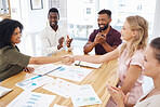 Female coworkers shaking hands while their colleagues clap hands. Group of diverse businesspeople having a meeting at a table in an office. Business professionals talking and planning together at work