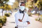 One African American businessman standing alone in a city with his arms folded and wearing a Covid face mask during a morning commute to an office. Confident black man during pandemic staying safe