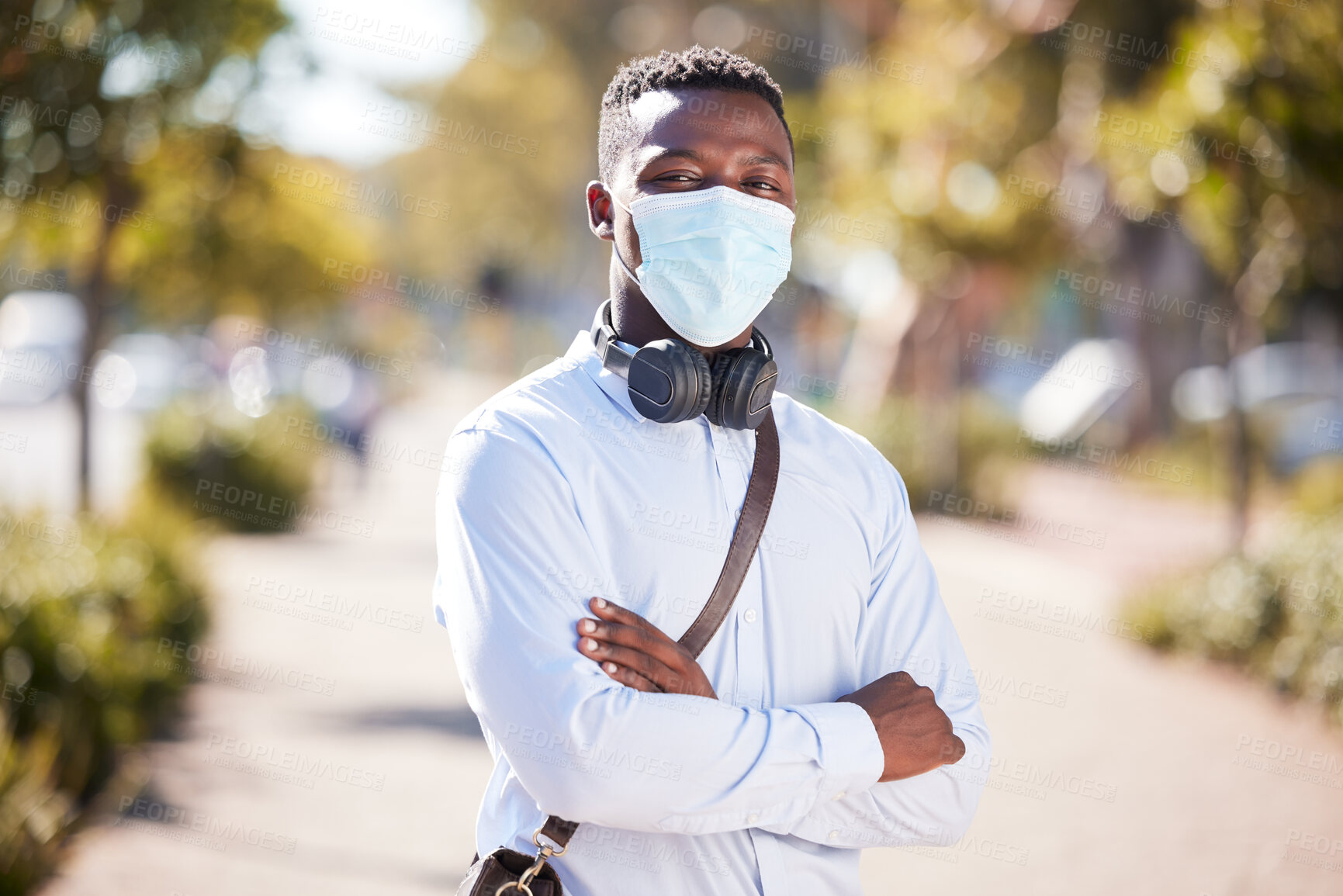 Buy stock photo One African American businessman standing alone in a city with his arms folded and wearing a Covid face mask during a morning commute to an office. Confident black man during pandemic staying safe