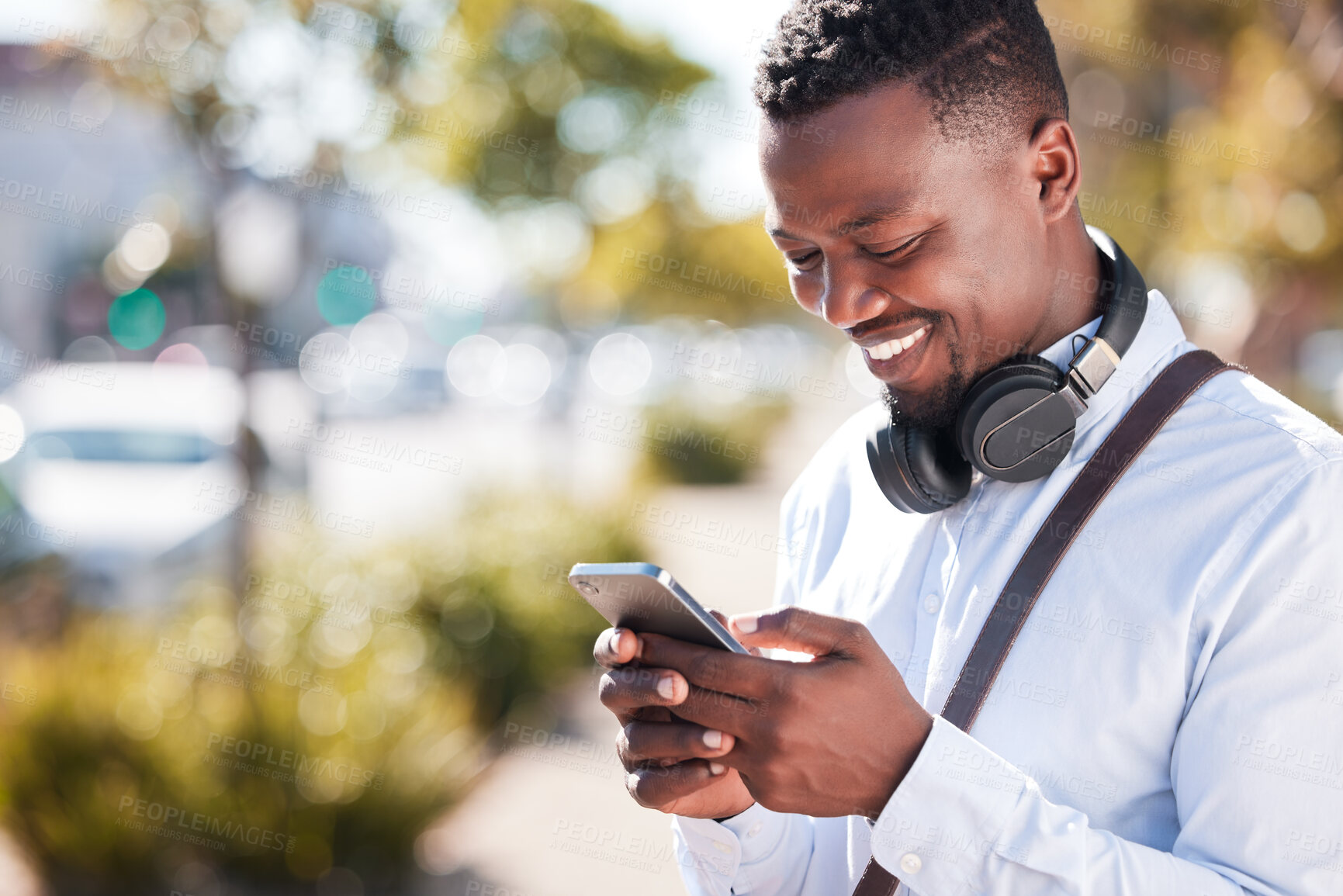 Buy stock photo Happy African American man wearing headphones and texting on a smartphone while out in the city on daily commute. Black male smiling while checking social media and standing outside