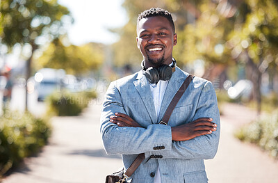 One african american man wearing a sling bag while standing with his arms crossed out in the city. Portrait of a happy handsome black male with wireless headphones around his neck on his work commute