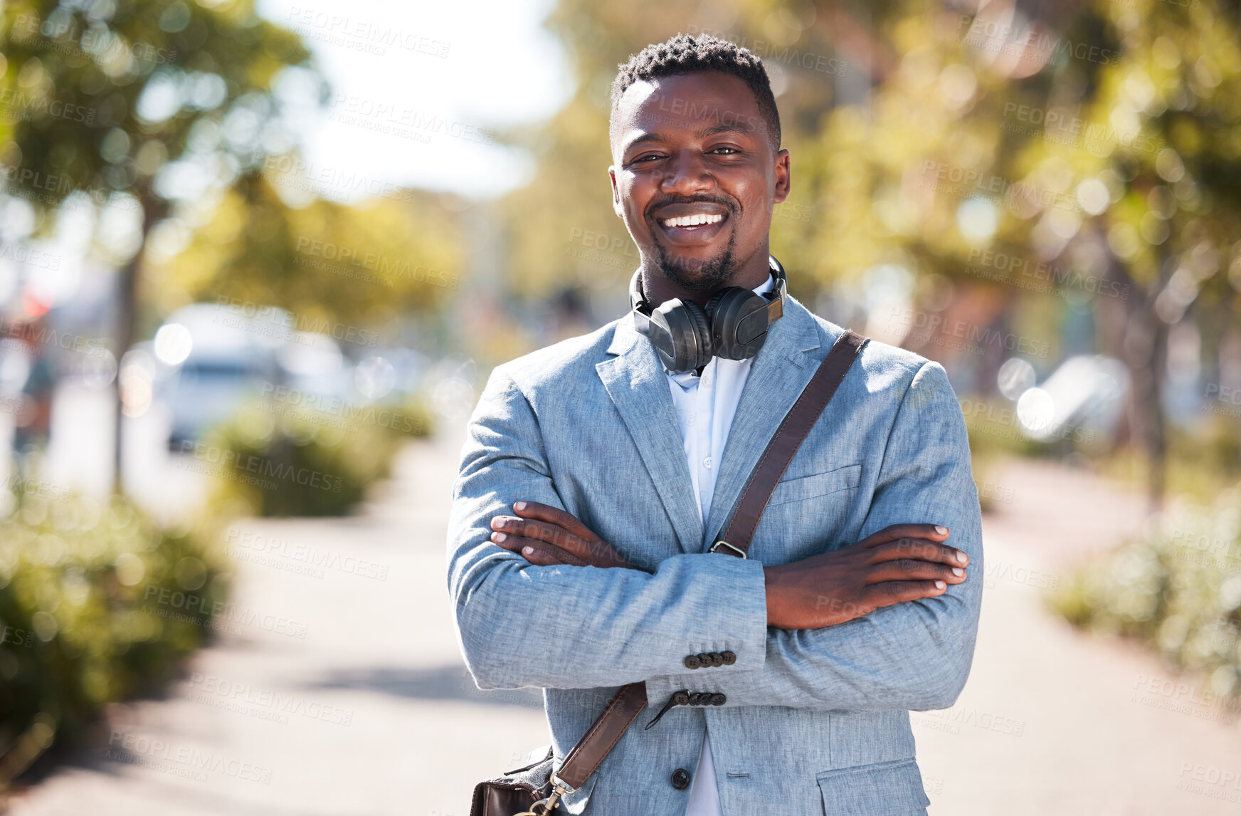 Buy stock photo One african american man wearing a sling bag while standing with his arms crossed out in the city. Portrait of a happy handsome black male with wireless headphones around his neck on his work commute