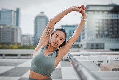 Portrait of a young mixed race female athlete looking serious while stretching her arms before her run in the city. Young woman warming up her muscles while during exercise against a urban background