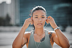 Young serious looking mixed race female athlete wearing earphones and listening to music while standing outside in the city. Positive, focused, workout. Taking a break