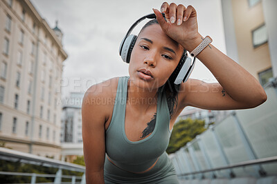 Buy stock photo Portrait of a young mixed race female athlete wearing wireless headphones and listening to music while standing outside in the city wiping her sweaty forehead and tired from a workout. Determined to get a fit and toned body