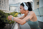 Young serious mixed race hispanic female athlete looking thoughtful wearing headphones and listening to music while standing on a bridge outside In the city. Taking a break from her excising. Determined to achieve my goal