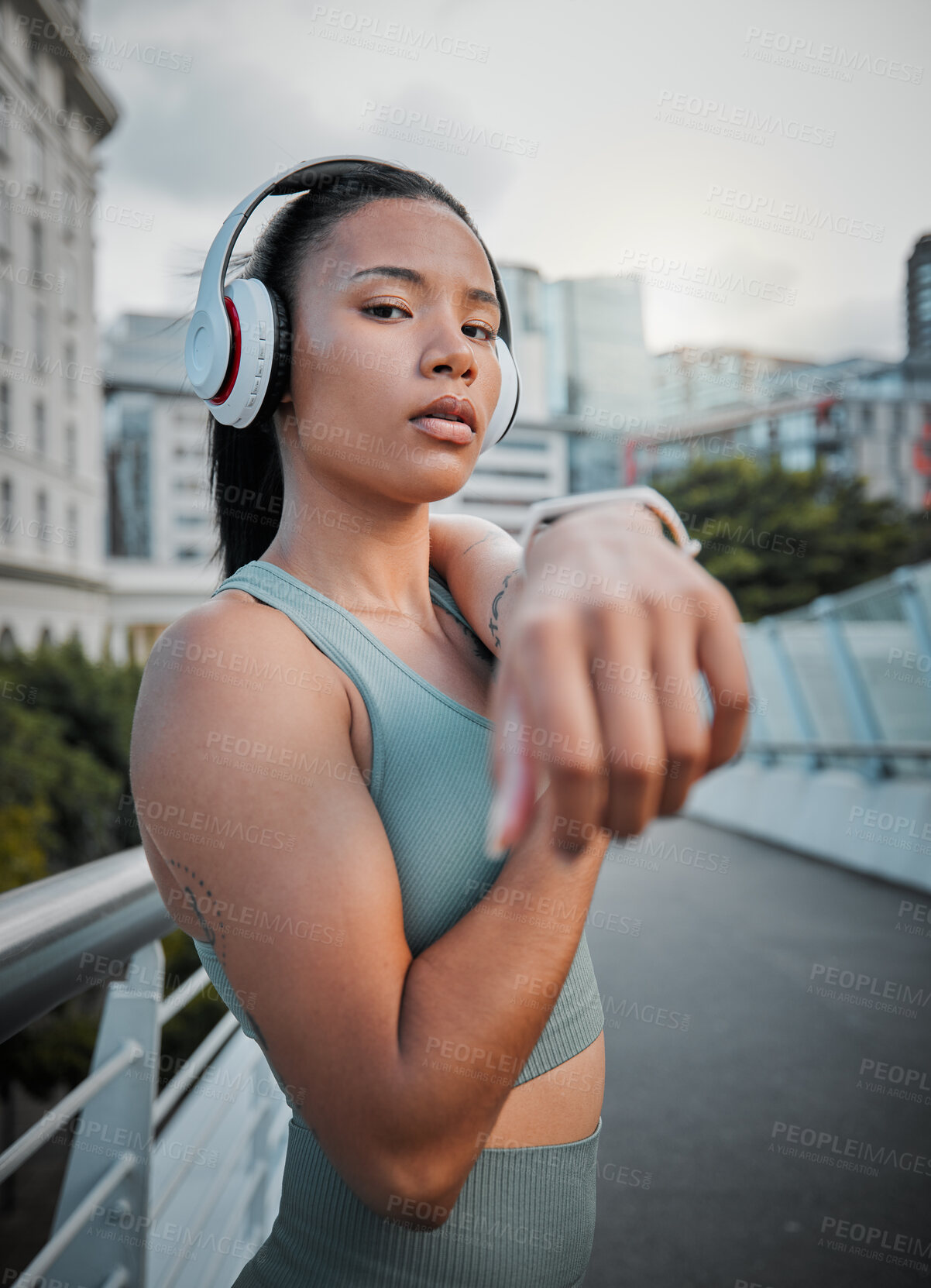 Buy stock photo Young mixed race female athlete wearing headphones and listening to music while stretching her muscles before a run outside in the city. Warming up before exercising to improve her health and lifestyle