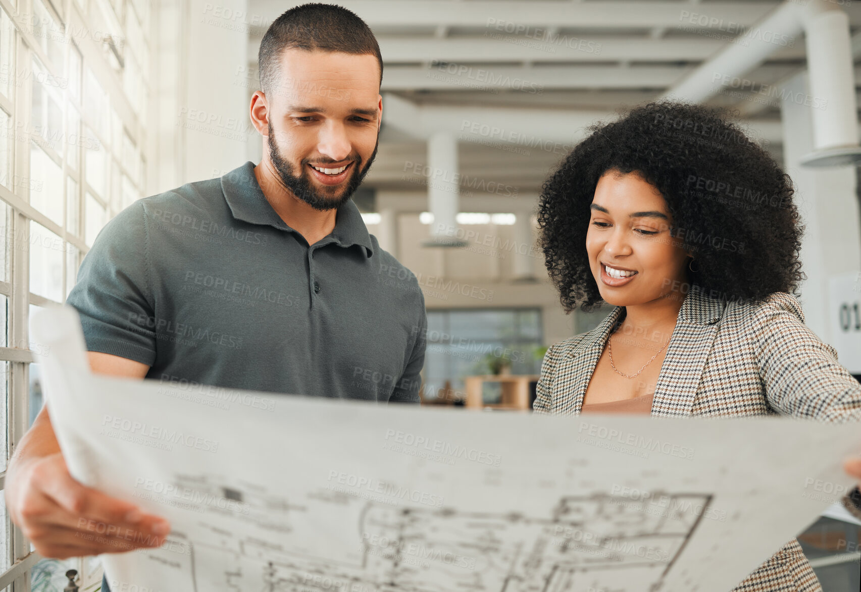 Buy stock photo Young coworkers looking at a blueprint. Two businesspeople collaborate on a building plan. Design creatives working together in a modern office. Happy colleagues plan a building project together